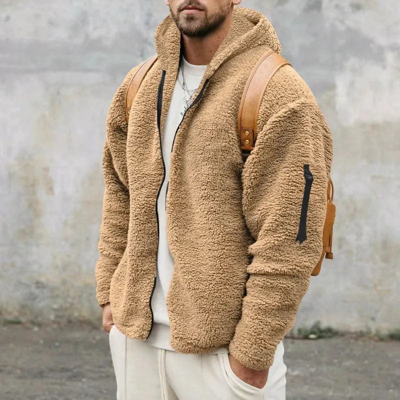 Mann in beiger Teddy-Fleecejacke mit Kapuze und braunem Rucksack vor grauer Wand. Modetrend, Herrenmode, Winterjacke, Streetwear.
