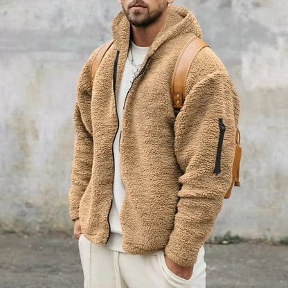 Mann in beiger Teddy-Fleecejacke mit Kapuze und braunem Rucksack vor grauer Wand. Modetrend, Herrenmode, Winterjacke, Streetwear.