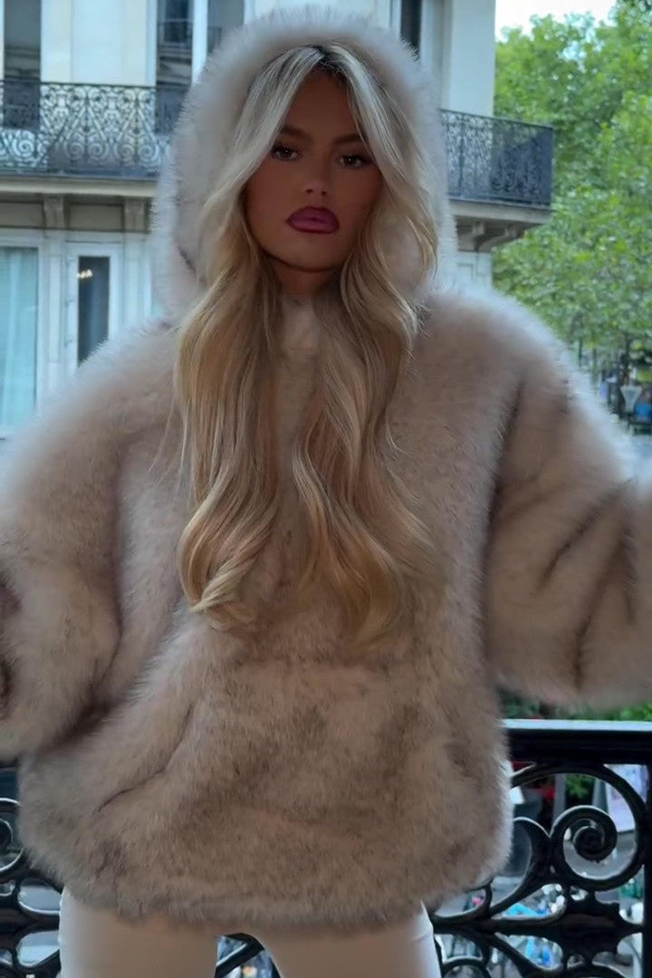 Blonde Frau in luxuriösem, flauschigem Pelzmantel mit Kapuze, steht auf Balkon in städtischer Umgebung. Elegante Mode, Herbst/Winter Look.