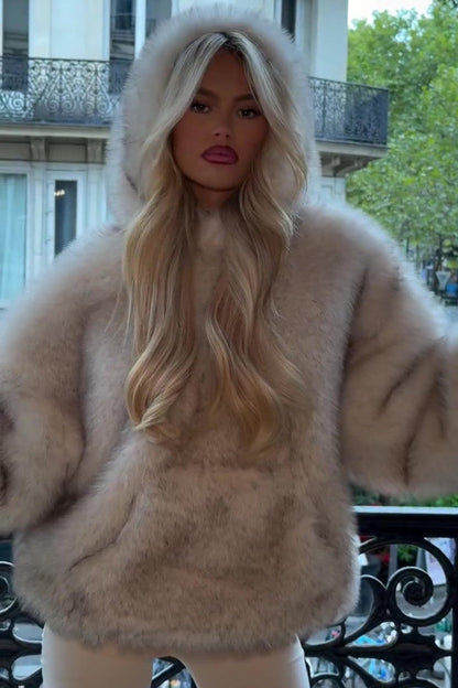 Blonde Frau in luxuriösem, flauschigem Pelzmantel mit Kapuze, steht auf Balkon in städtischer Umgebung. Elegante Mode, Herbst/Winter Look.