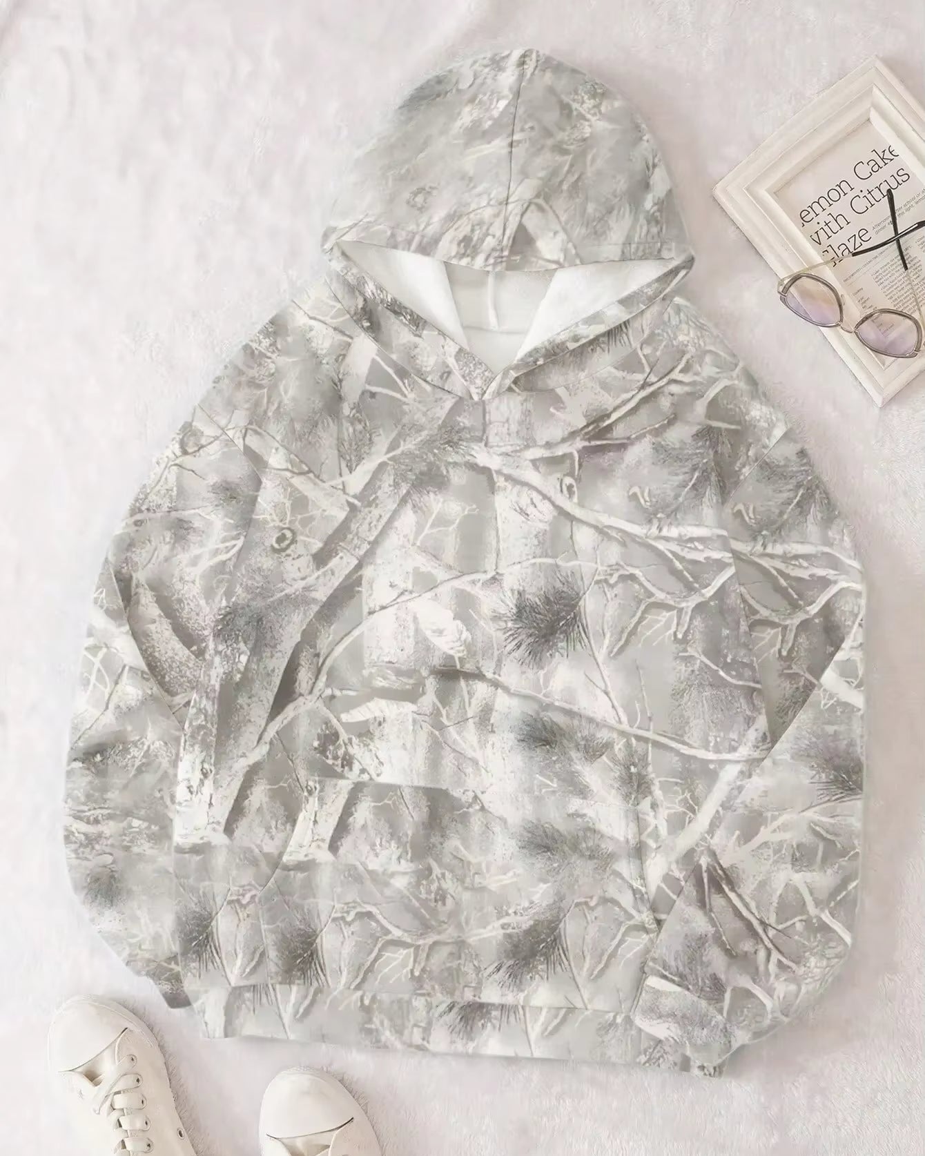 Grauer Camouflage-Hoodie mit Waldmuster auf weißem Hintergrund, daneben eine Brille und ein Buch. Bequeme Freizeitmode für Outdoor-Aktivitäten.