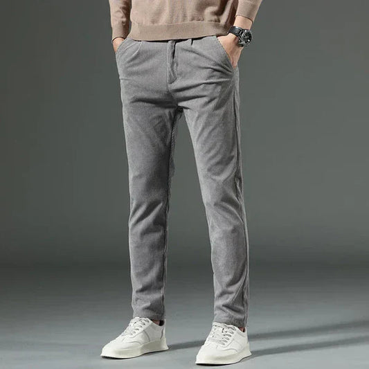 Graue Cordhose für Herren, kombiniert mit weißen Sneakers und beigem Pullover. Modischer Freizeitlook, ideal für Herbst und Winter.