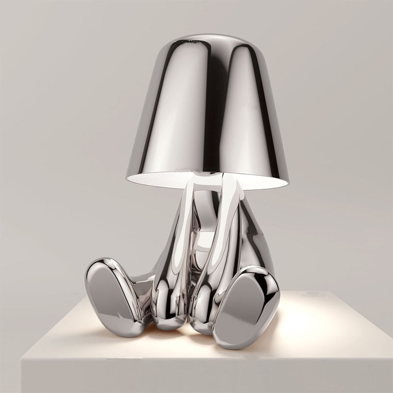 Silberne Designer-Tischlampe in Form einer sitzenden Figur, modernes minimalistisches Design, glänzende Metalloberfläche, stilvolle Wohnkultur.