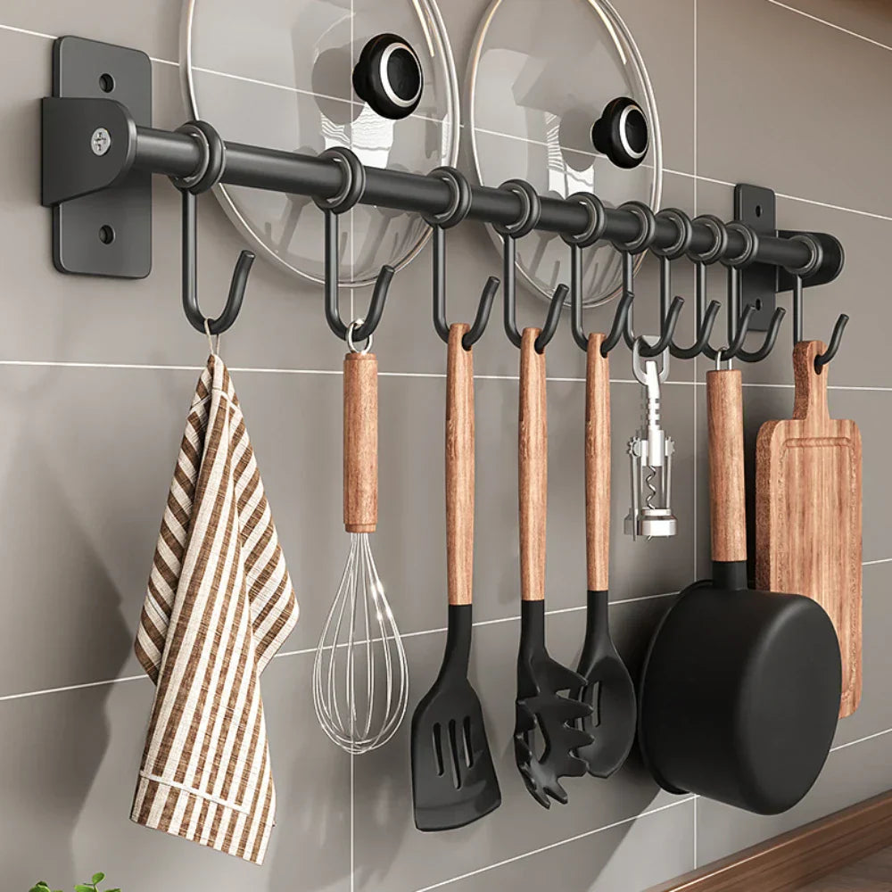 Schwarze Küchenleiste mit Haken, an denen Kochutensilien aus Holz und Metall hängen, inklusive Pfanne, Schneebesen, Schöpfkelle und Geschirrtuch.