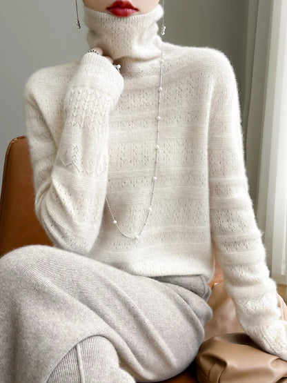 Frau in cremefarbenem Strickpullover mit Rollkragen, eleganter Halskette und rotem Lippenstift, sitzt auf braunem Stuhl. Modetrend Herbst/Winter.