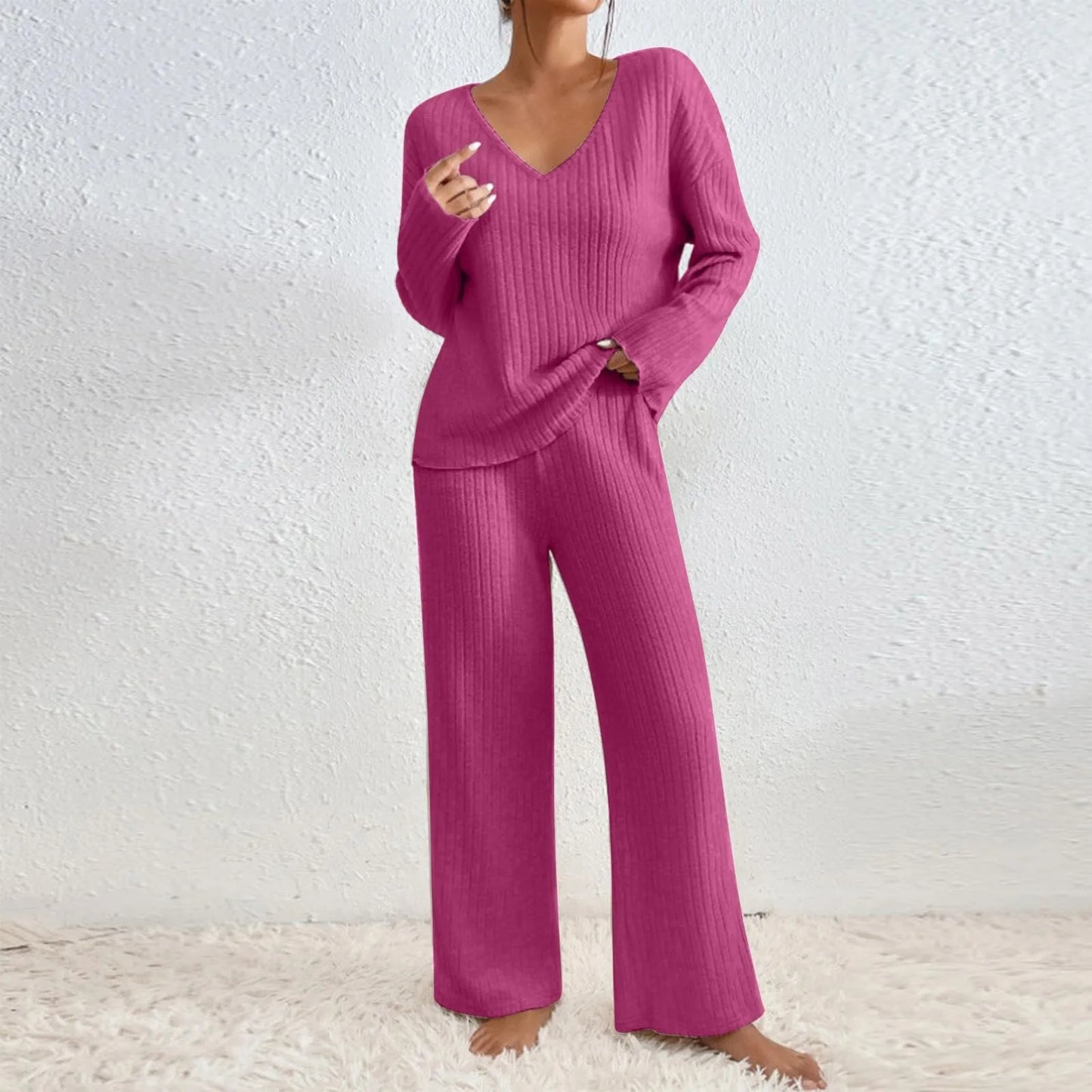 Frau in pinkem Strick-Loungewear-Set, bestehend aus langärmligem Oberteil und weiter Hose, steht barfuß auf einem weißen Teppich vor weißer Wand.