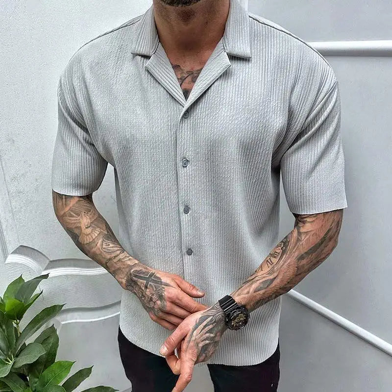 Mann in grauem Kurzarmhemd mit Knopfleiste, tätowierten Armen und schwarzer Uhr, steht vor weißer Wand. Herrenmode, Sommeroutfit, stilvoll.
