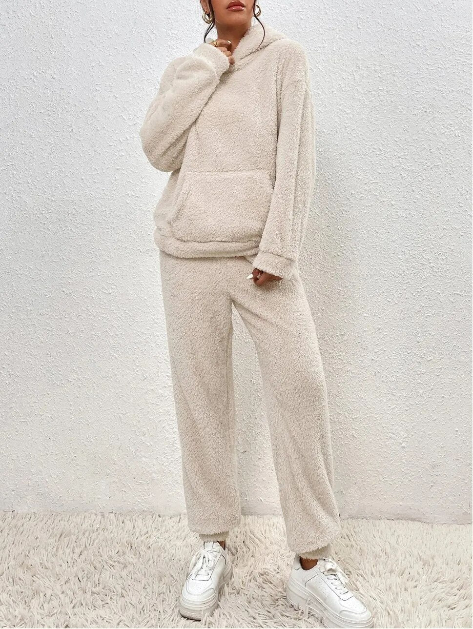 Frau in beigem Fleece-Loungewear-Set mit Kapuze und Kängurutasche, steht vor weißer Wand auf Teppich, trägt weiße Sneaker. Gemütliche Mode.