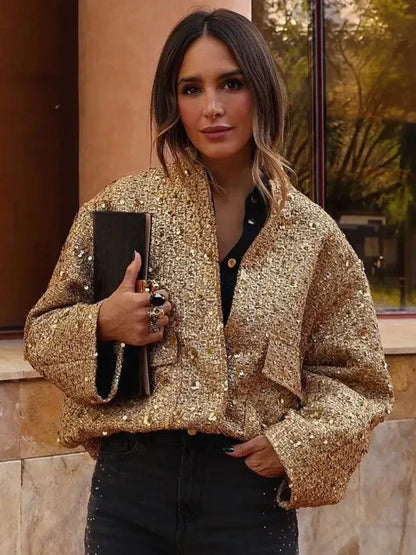 Frau in goldener Paillettenjacke, hält schwarze Clutch, steht vor Gebäude. Modetrend, glamouröser Look, stilvolle Abendgarderobe.