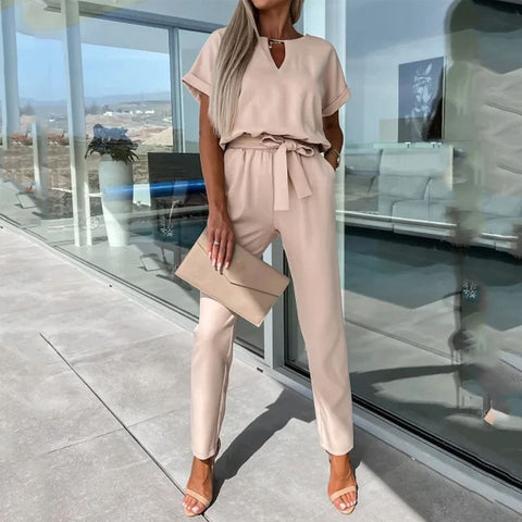 Frau in eleganter, beigefarbener Jumpsuit mit Gürtel, hält eine passende Clutch. Sommermode, stilvoll, modern, draußen vor Glasfassade.