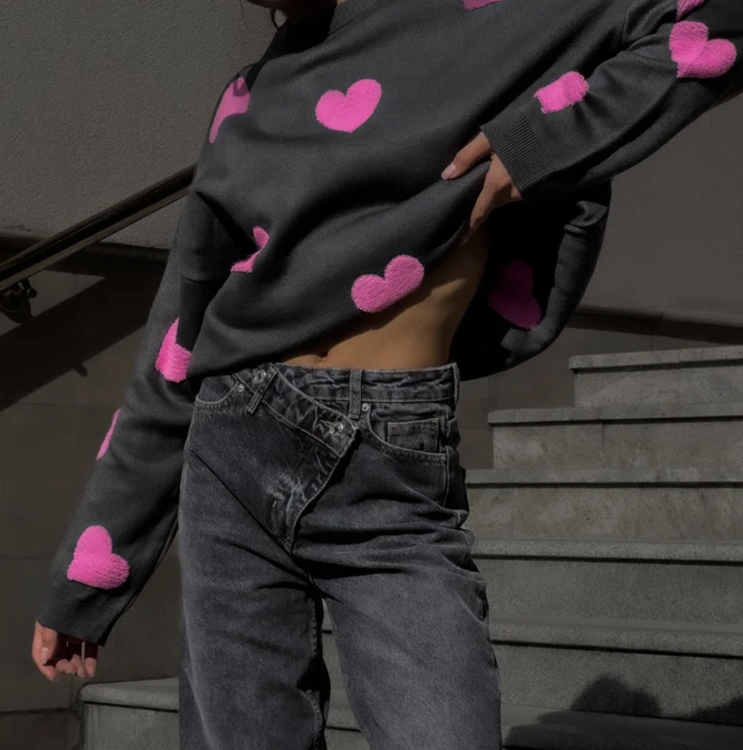 Grauer Pullover mit pinken Herzen, kombiniert mit hoch taillierten, überkreuzten Jeans. Modetrend, Streetstyle, Herbstmode.