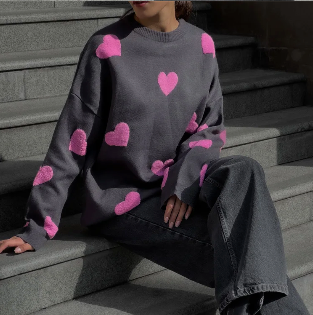 Grauer Pullover mit rosa Herzmustern, getragen von einer Person auf Treppen. Modetrend, lässiger Stil, Herbstmode, Damenbekleidung.