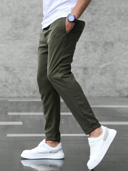 Mann in olivgrüner Chinohose und weißen Sneakers, seitlich stehend, mit Händen in den Taschen, vor grauer Wand. Moderne Herrenmode, lässiger Stil.