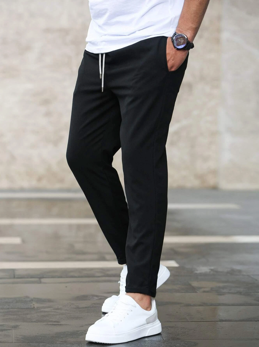 Mann in schwarzer Jogginghose und weißen Sneakers, trägt weißes T-Shirt, steht auf grauem Boden. Lässige Herrenmode, sportlicher Look.