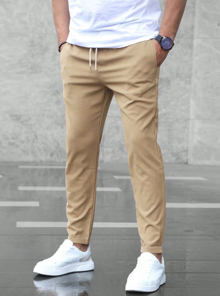 Mann in beiger Chinohose und weißen Sneakers vor grauem Hintergrund. Lässiger Herrenstil, moderne Mode, Freizeitlook.