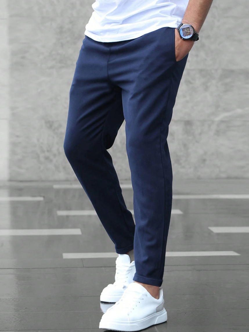 Mann in blauer Chino-Hose und weißen Sneakers vor grauem Hintergrund. Moderne Herrenmode, lässiger Stil, trendige Freizeitkleidung.