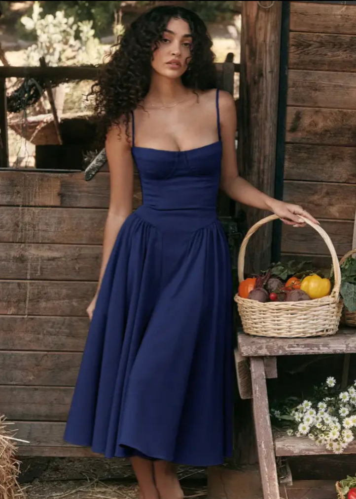 Frau in blauem Sommerkleid steht vor Holzhütte, hält Korb mit Gemüse und Kürbis. Ländliche Mode, Herbsternte, rustikaler Stil.