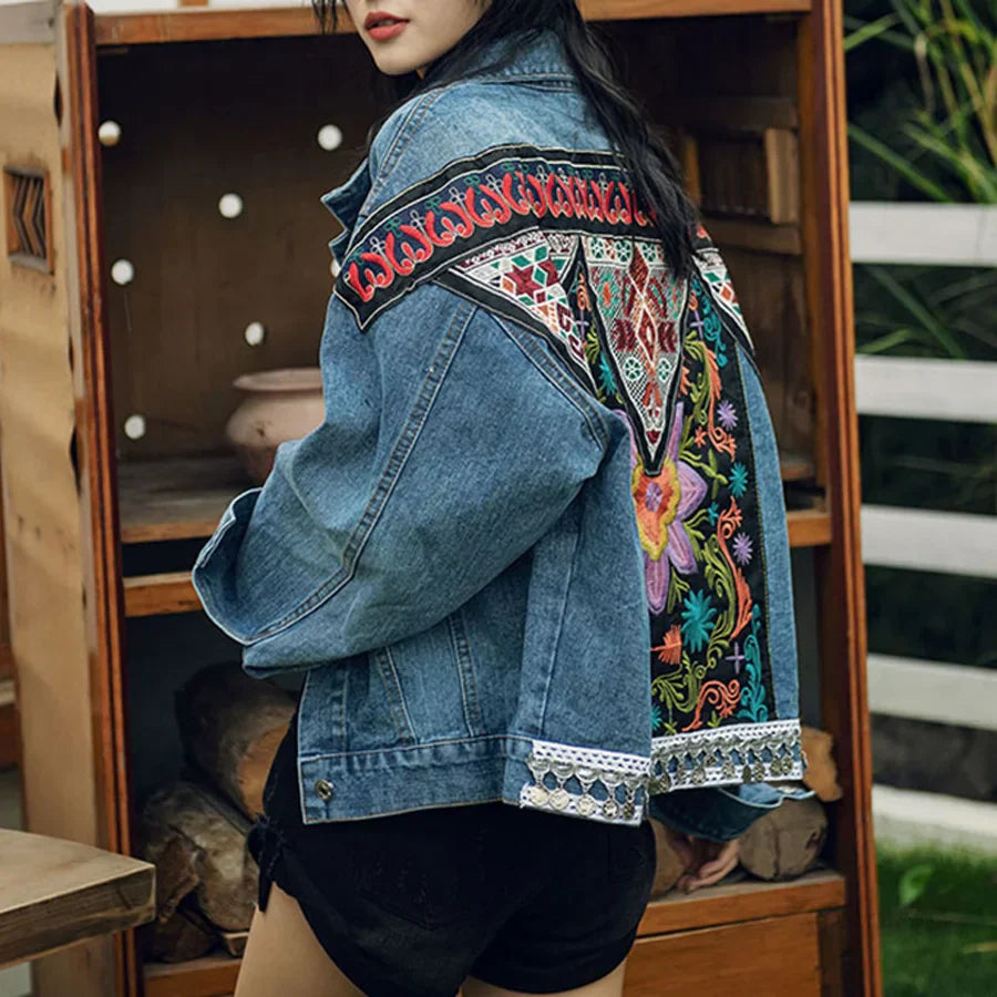 Frau in bestickter Jeansjacke mit buntem Blumenmuster, steht vor Holzregal. Modetrend, Damenmode, Streetstyle, Denim, Stickerei.