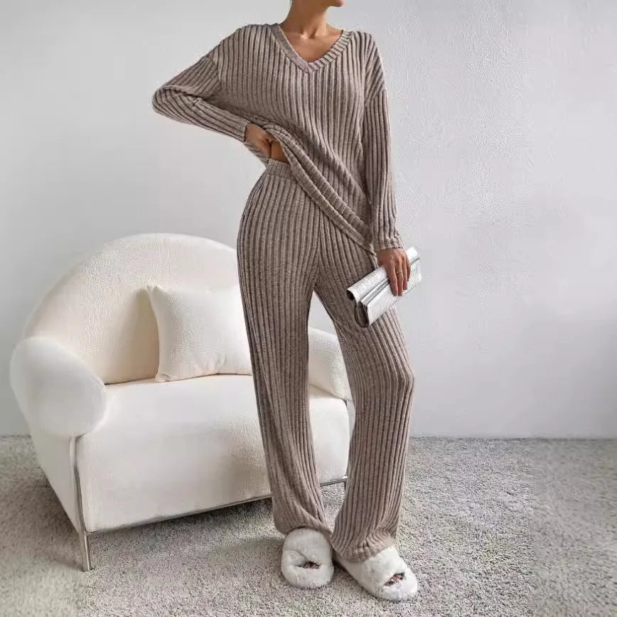 Frau in beigem Strick-Loungewear-Set mit V-Ausschnitt, hält silberne Clutch, steht neben weißem Sessel auf grauem Teppich. Gemütliche Mode.