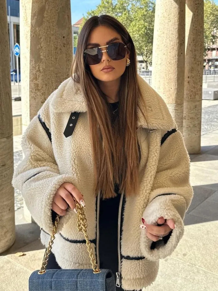 Frau mit langer brauner Haaren trägt übergroße beige Teddyjacke, schwarze Sonnenbrille und hält blaue Handtasche mit goldener Kette.