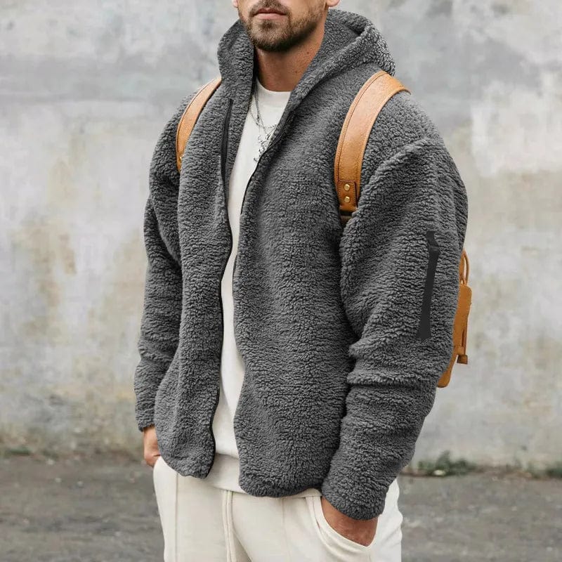 Mann in grauer Teddyfleece-Jacke mit Kapuze und braunem Rucksack vor grauer Wand. Modischer Winterlook, lässiger Stil.