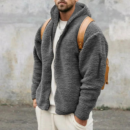 Mann in grauer Teddyfleece-Jacke mit Kapuze und braunem Rucksack vor grauer Wand. Modischer Winterlook, lässiger Stil.