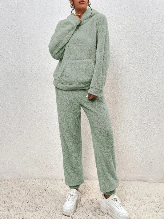 Frau in grünem Fleece-Loungewear-Set mit Kapuze und Kängurutasche, steht auf Teppich vor weißer Wand, trägt weiße Sneaker.