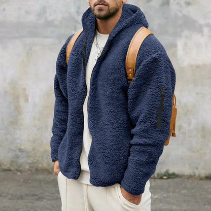 Mann in blauer Fleecejacke mit Kapuze und braunem Rucksack vor grauer Wand. Modetrend, Herrenbekleidung, Wintermode, Outdoor-Look.