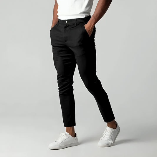 Mann in schwarzer Hose und weißen Sneakers, Hände in den Taschen, vor grauem Hintergrund. Moderne Herrenmode, lässiger Stil.