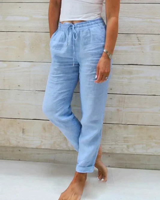 Frau trägt hellblaue Leinenhose mit Kordelzug, steht barfuß vor Holzwand. Sommermode, lässiger Stil, bequeme Damenbekleidung.