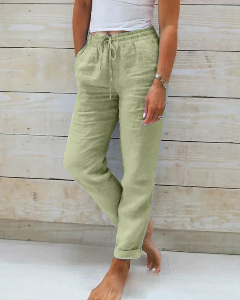 Frau trägt grüne Leinenhose mit Kordelzug, steht barfuß vor Holzwand. Sommermode, lässiger Stil, bequeme Damenbekleidung.