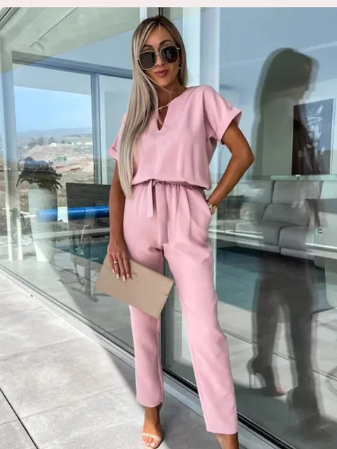 Frau in rosa Jumpsuit mit Sonnenbrille, steht auf Terrasse vor Glaswand, hält beigen Umschlag. Elegante Sommermode, stilvolle Damenbekleidung.