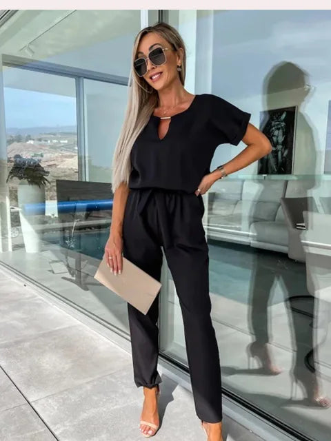Frau in eleganter schwarzer Kleidung mit Sonnenbrille und Clutch steht auf moderner Terrasse. Stilvolle Mode, Sommeroutfit, urbane Eleganz.