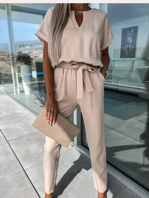 Frau in beigem Jumpsuit mit Gürtel, hält eine passende Clutch. Stilvolle Sommermode, elegantes Outfit, moderne Damenbekleidung.