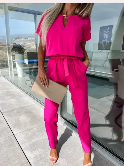 Frau in leuchtend pinkem Jumpsuit mit beiger Clutch, steht in modernem, lichtdurchflutetem Raum. Elegante Mode, Sommeroutfit, stilvolle Kleidung.