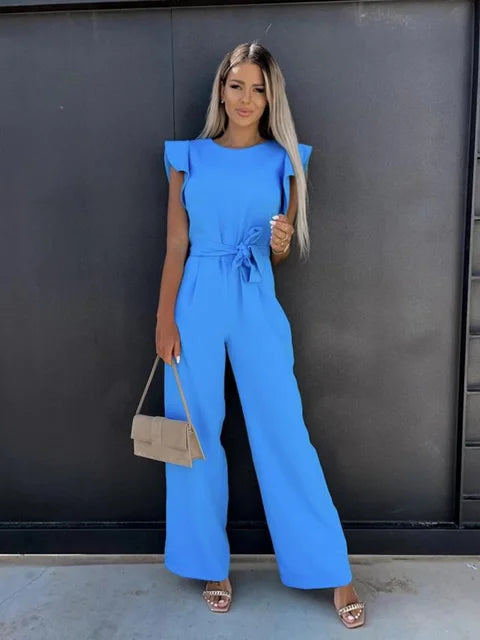 Frau in blauem Jumpsuit mit Gürtel, hält beige Handtasche, steht vor schwarzer Wand. Elegante Mode, Sommeroutfit, Damenbekleidung.