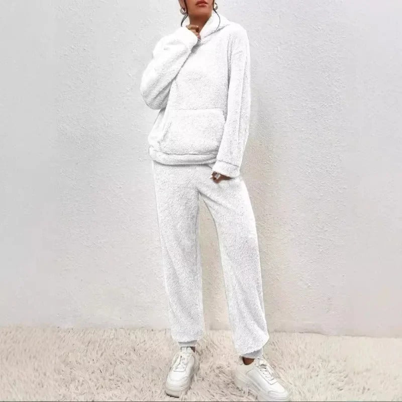 Frau in weißem Fleece-Hoodie und Jogginghose, lässiger Look, gemütliche Wintermode, weiße Sneaker, minimalistische Wand, modische Freizeitkleidung.