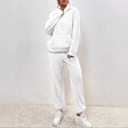 Frau in weißem Fleece-Hoodie und Jogginghose, lässiger Look, gemütliche Wintermode, weiße Sneaker, minimalistische Wand, modische Freizeitkleidung.