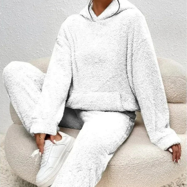 Frau in weißem, flauschigem Fleece-Hoodie und Jogginghose sitzt auf beigem Sessel, trägt weiße Sneaker. Bequeme, gemütliche Loungewear.