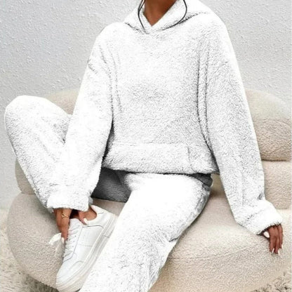 Frau in weißem, flauschigem Fleece-Hoodie und Jogginghose sitzt auf beigem Sessel, trägt weiße Sneaker. Bequeme, gemütliche Loungewear.