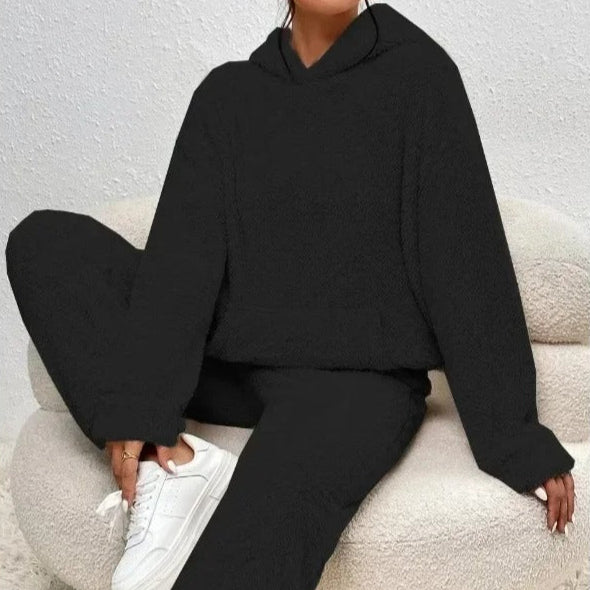 Frau in schwarzem Hoodie und Jogginghose sitzt auf beigem Sessel, trägt weiße Sneaker. Bequeme Freizeitmode, gemütlicher Look.