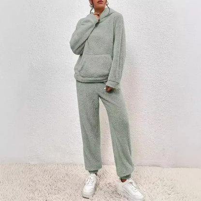 Dame in grauem Fleece-Loungewear-Set mit Kapuze und Kängurutasche, kombiniert mit weißen Sneakern, steht vor weißer Wand auf Teppich.