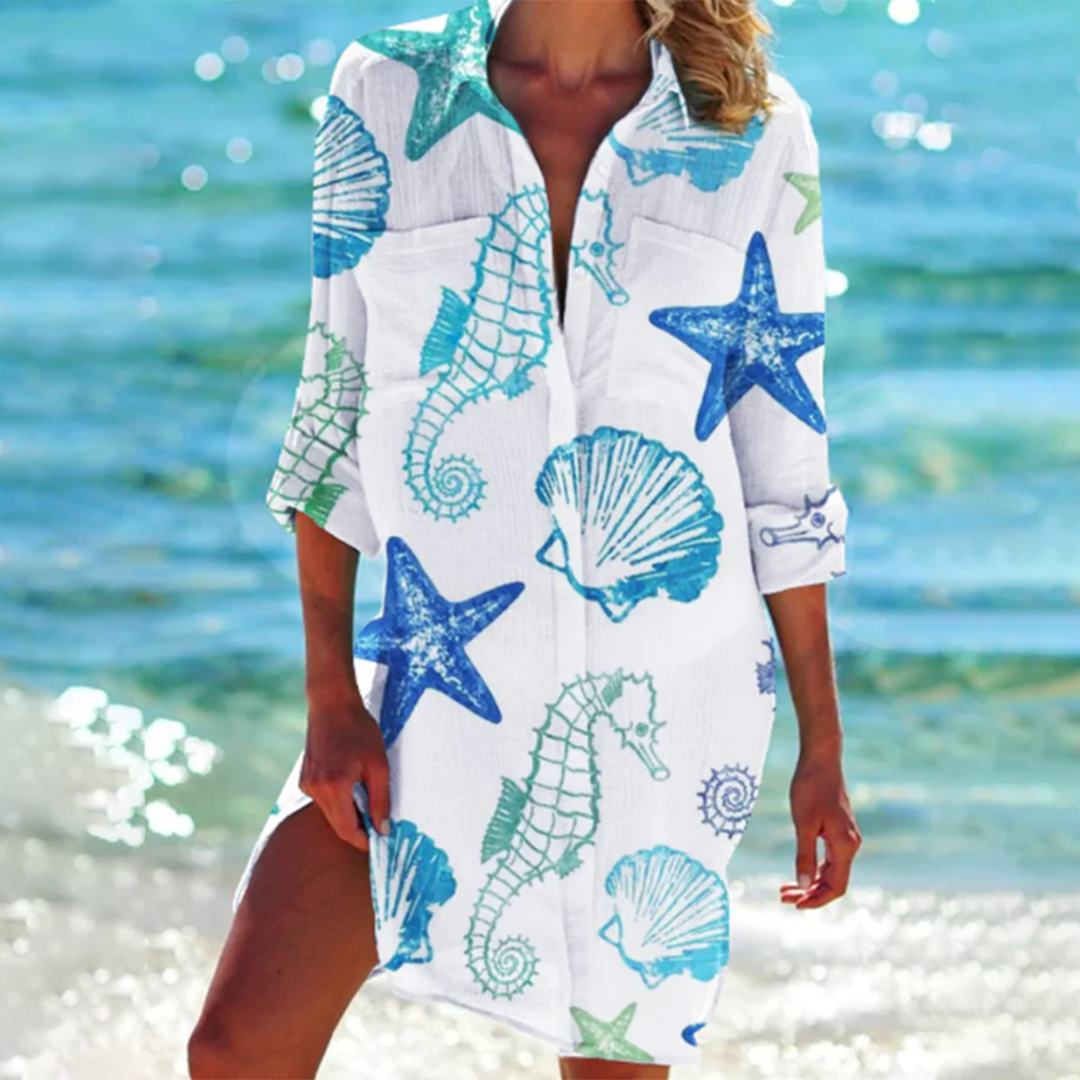 Frau am Strand in weißem Strandkleid mit blauen Seesternen, Seepferdchen und Muscheln. Sommermode, Strandkleidung, maritimes Design.