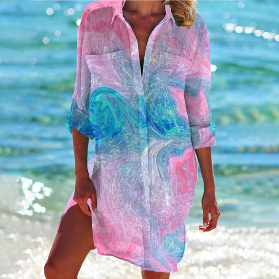 Frau am Strand in buntem, marmoriertem Strandkleid, türkis-blaues Meer im Hintergrund, Sommermode, Strandoutfit, Urlaubskleidung.