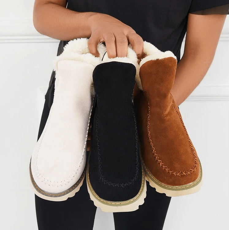 Damen hält drei Paar Winterstiefel aus Wildleder in Weiß, Schwarz und Braun mit Fellfutter. Bequeme, warme Schuhe für kalte Tage.