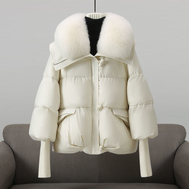 Weiße Daunenjacke mit großem Pelzkragen, Reißverschluss und Taschen, auf grauem Sofa drapiert. Wintermode, warme Damenbekleidung, stilvoll.