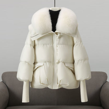 Weiße Daunenjacke mit großem Pelzkragen, Reißverschluss und Taschen, auf grauem Sofa drapiert. Wintermode, warme Damenbekleidung, stilvoll.