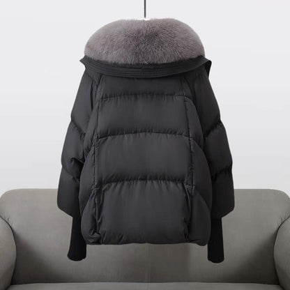 Schwarze Daunenjacke mit grauem Pelzkragen, hängend über einem grauen Sofa. Wintermode, warme Oberbekleidung, stilvolle Damenjacke.