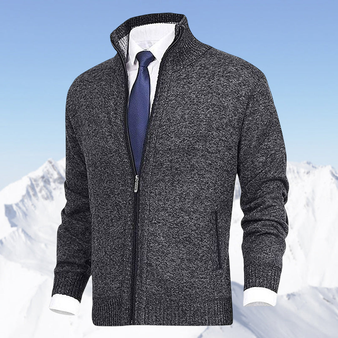 Grauer Herren-Strickjacke mit Reißverschluss, kombiniert mit weißem Hemd und blauer Krawatte, vor schneebedecktem Bergpanorama.