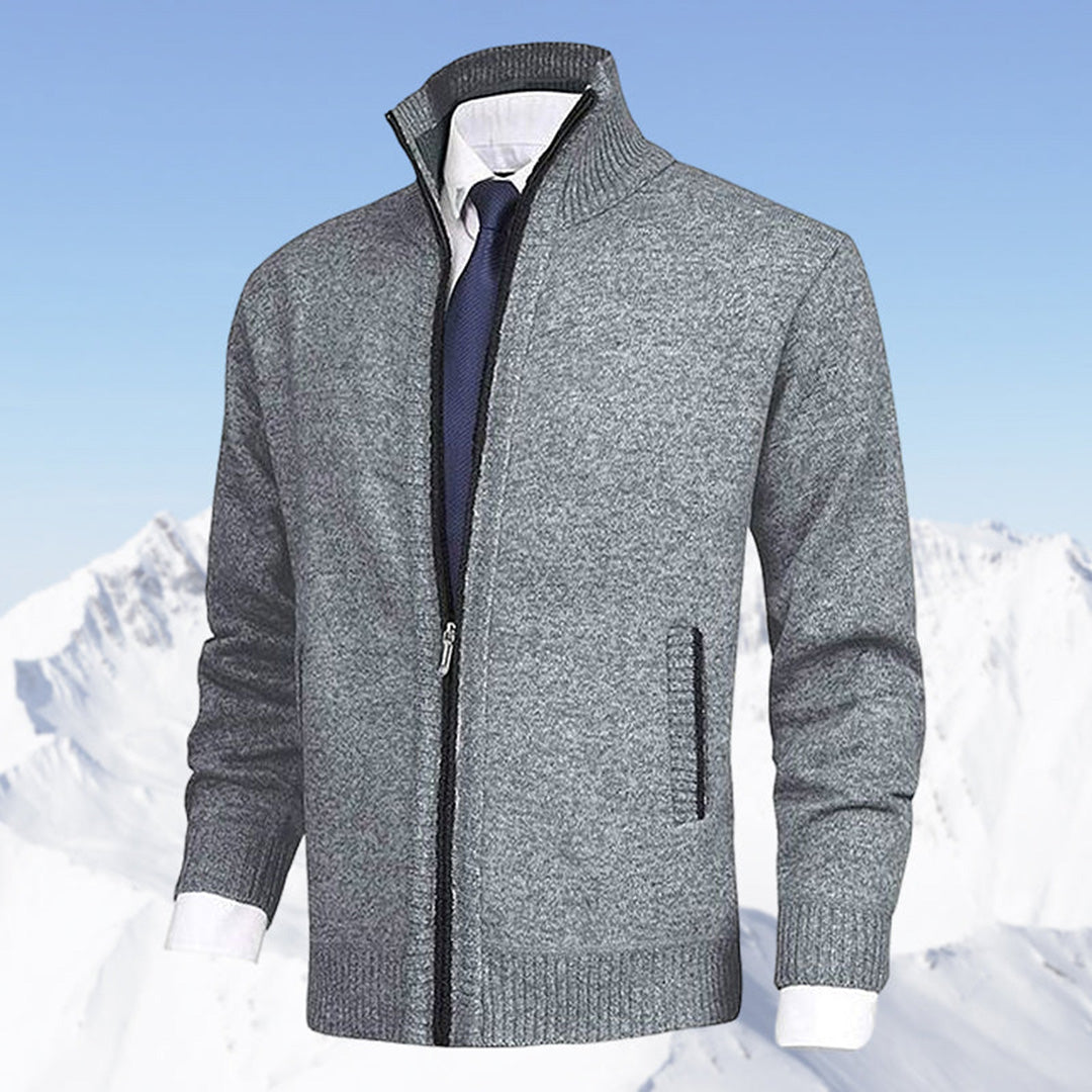 Grauer Herren-Strickjacke mit Reißverschluss, kombiniert mit weißem Hemd und blauer Krawatte, vor schneebedeckten Bergen. Elegante Wintermode.